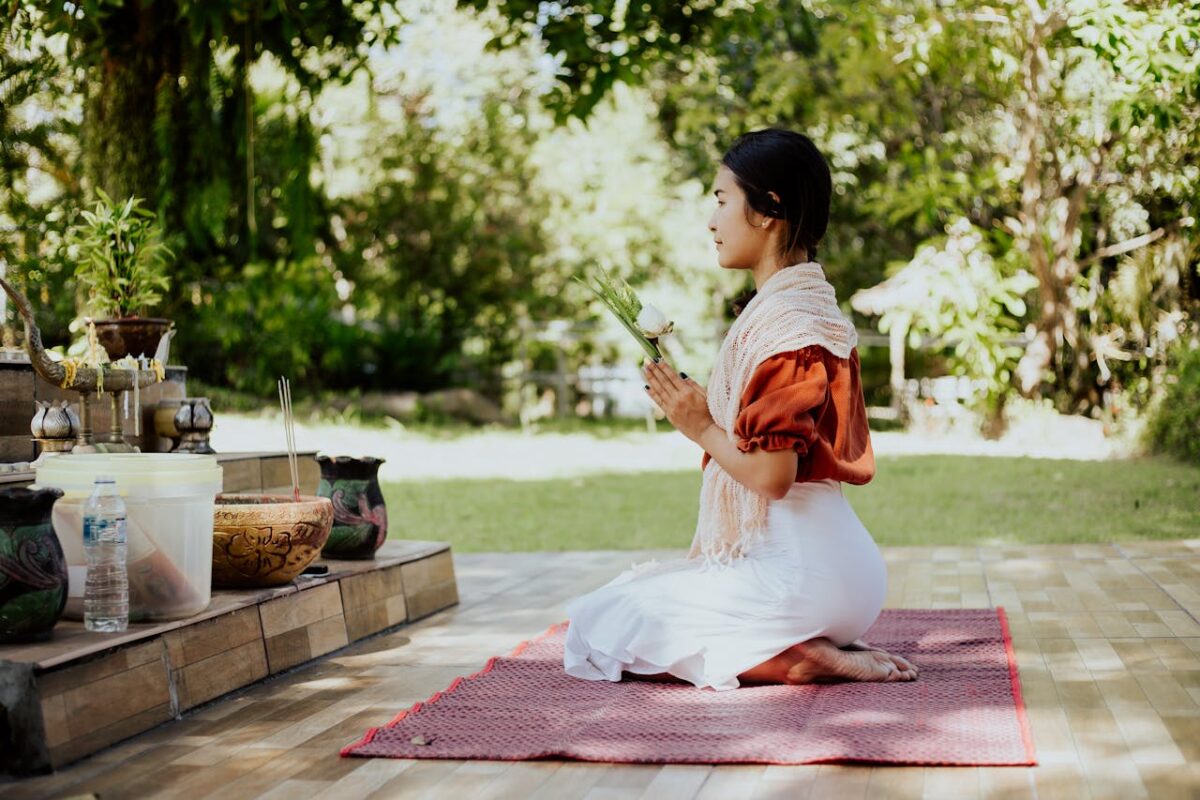 A woman kneels on a mat, praying outdoors in a serene garden setting.