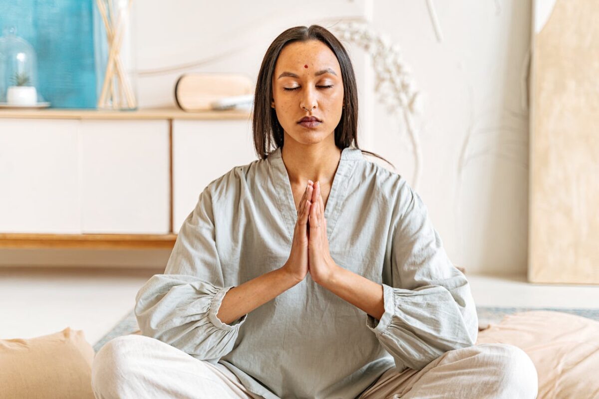 Woman meditating with eyes closed, hands together in peaceful indoor setting.